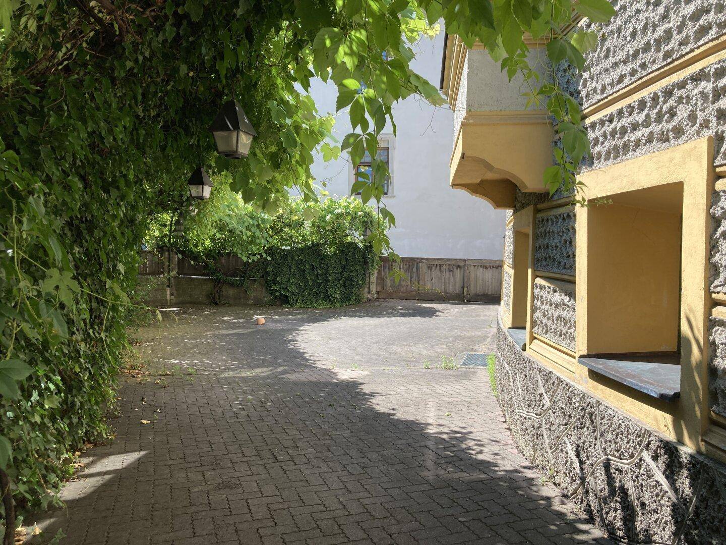 Gepflasterter Außenbereich mit üppigem Grün und der Fassade eines traditionellen Gebäudes im Sonnenlicht.