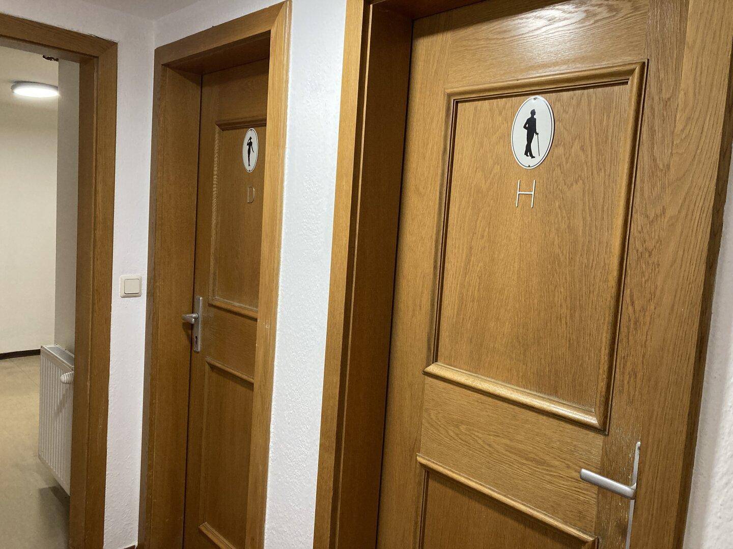Zwei Holztüren, eine mit Männersymbol, die zu den Toilettenräumen führen.