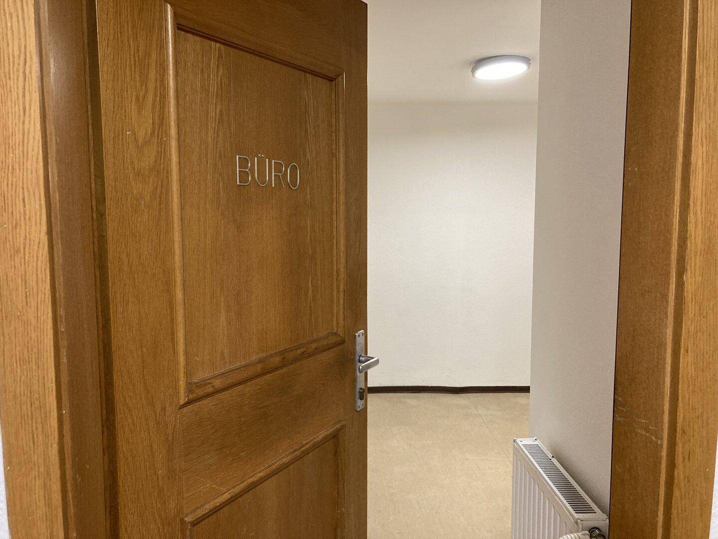 Offene Holztür mit der Aufschrift "BÜRO", die einen leeren Raum mit Heizkörper und hellem Boden zeigt.