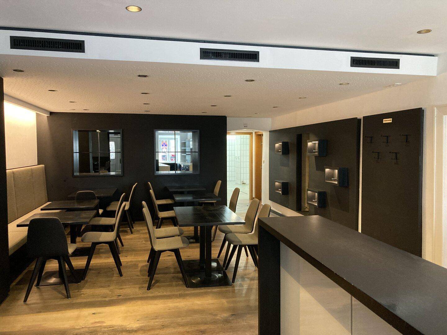 Moderner Restaurantbereich mit dunklen Wänden, Holzboden und Lüftungsschlitzen an der Decke.