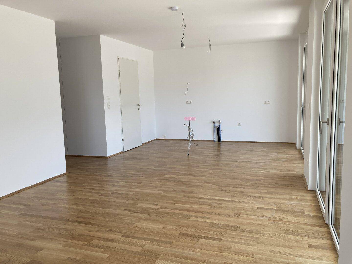 Heller, leerer Raum mit weißen Wänden, Holzfußboden und großen Fenstern, ideal für Wohn- oder Essbereich.
