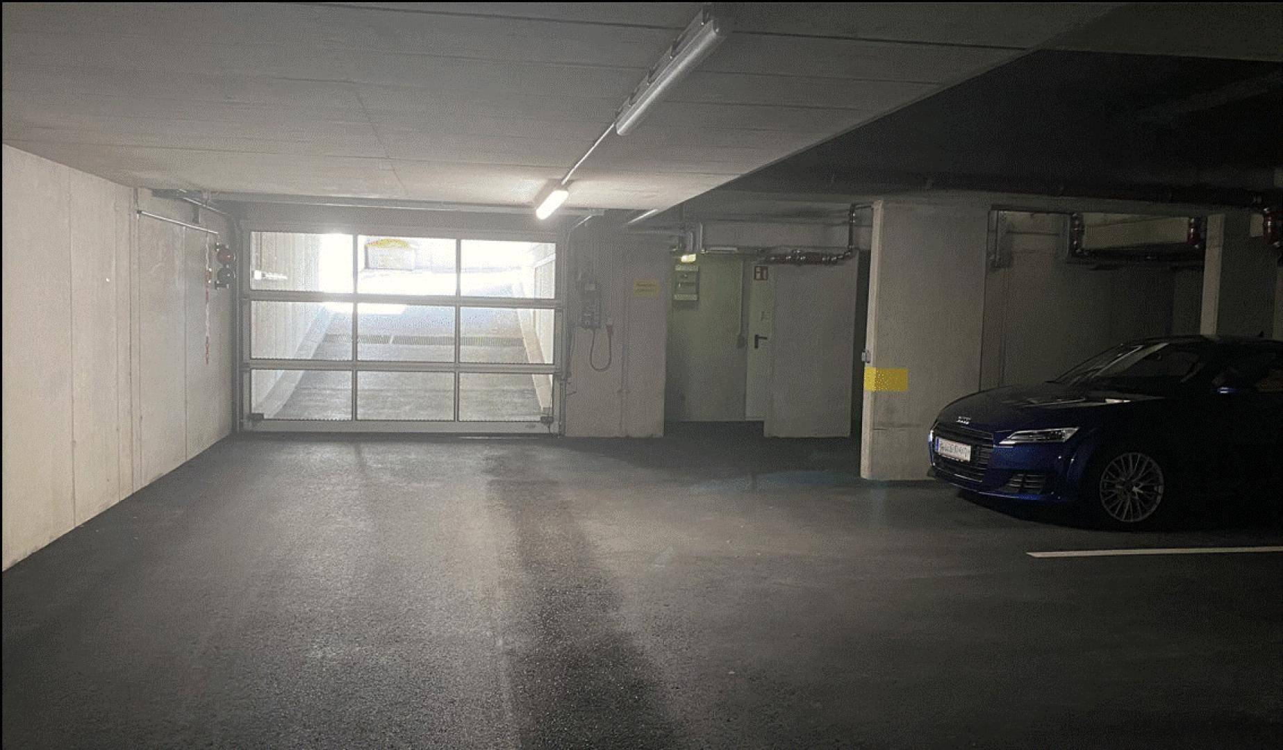 Geräumige Tiefgarage mit Einfahrtstor und einem geparkten blauen Auto, Betonstruktur und Beleuchtung.
