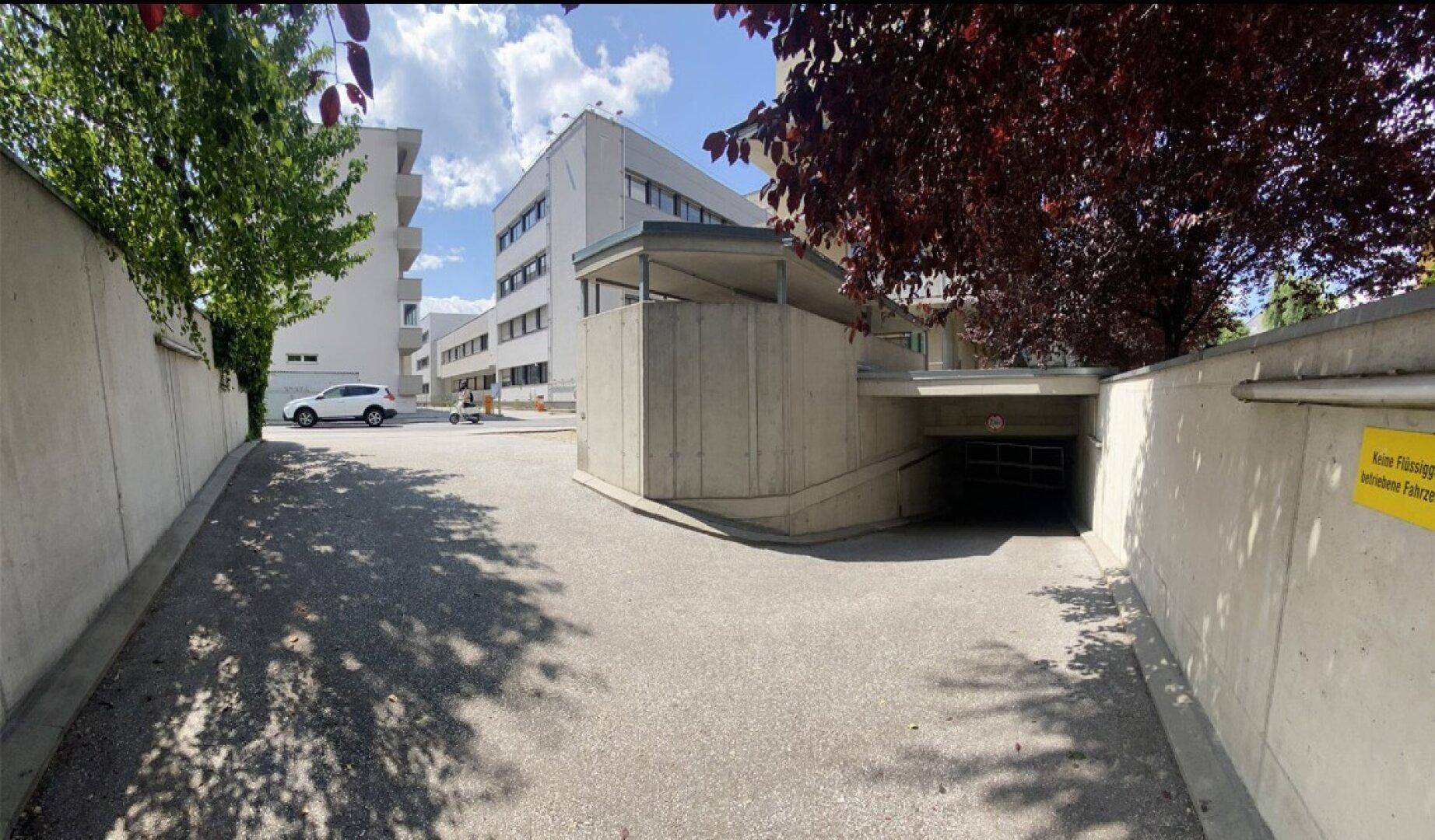 Außenzufahrt zu einer Tiefgarage mit Rampe, umgeben von modernen Gebäuden und Bäumen unter blauem Himmel.