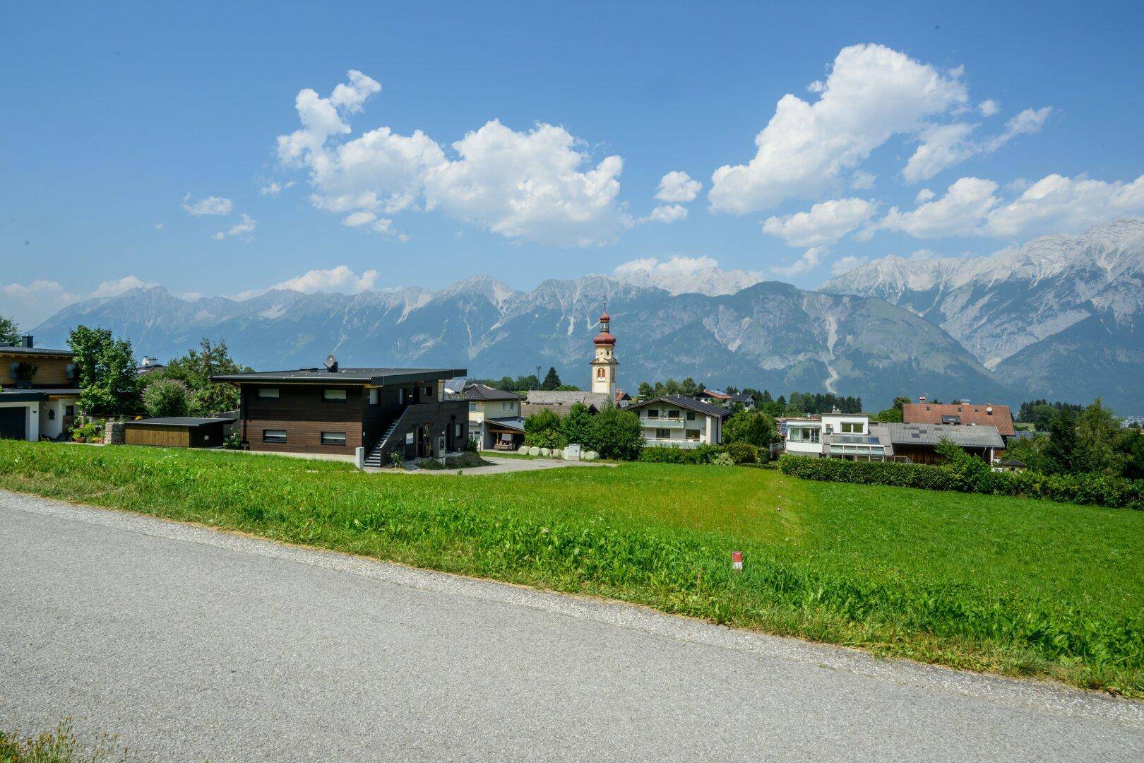 Malerischer Blick über grüne Felder auf ein Dorf mit Kirchturm und imposanter Bergkulisse.