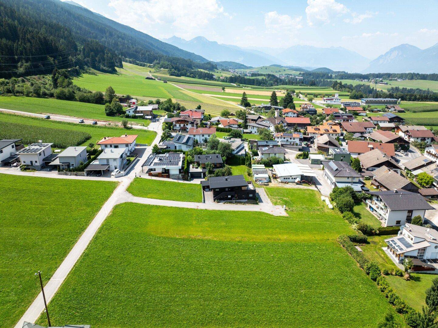 Panoramablick aus der Luft auf ein idyllisches Dorf, umgeben von grünen Feldern und bewaldeten Hügeln.