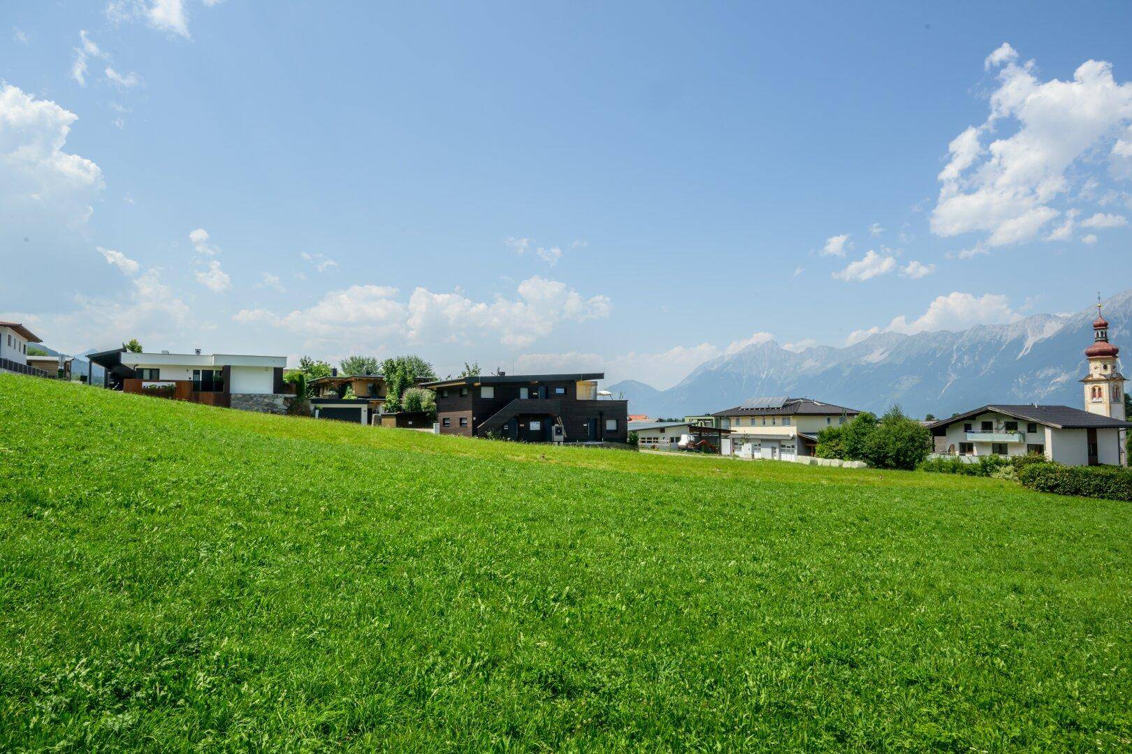 Sanft abfallende grüne Wiese mit Blick auf Häuser, einen Kirchturm und die majestätische Bergkette.