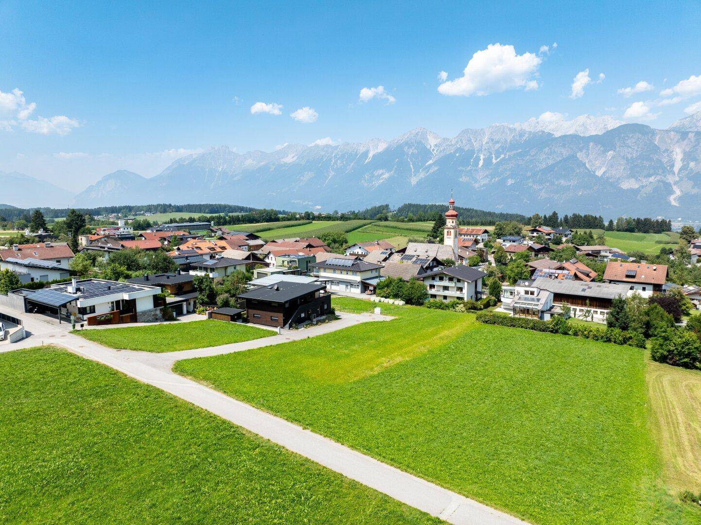 Weitläufige Luftaufnahme eines charmanten Dorfes, eingebettet in grüne Wiesen mit beeindruckendem Bergpanorama.