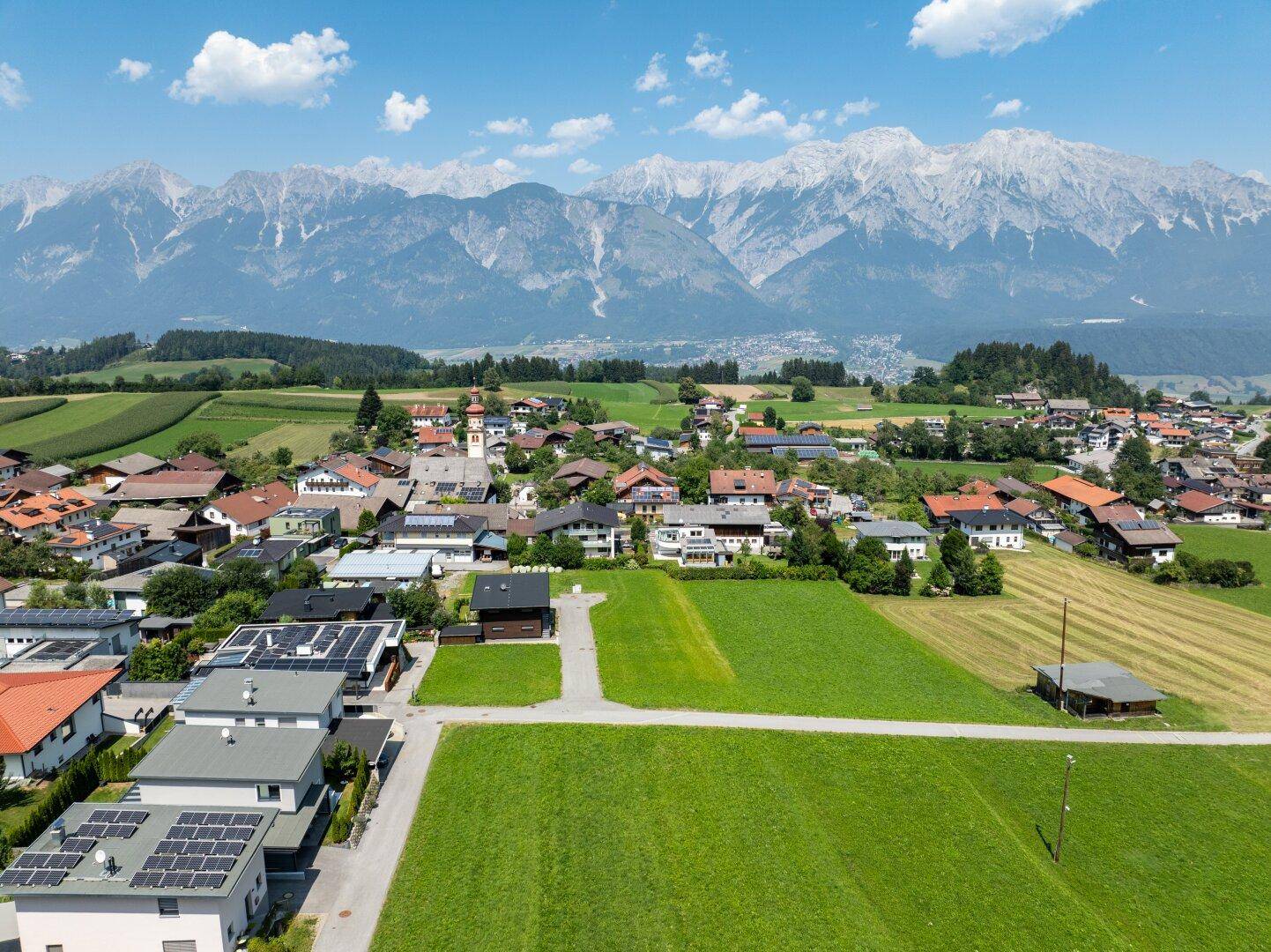 Panoramische Luftaufnahme eines Dorfes, umgeben von weiten grünen Feldern und einer beeindruckenden Bergkulisse.
