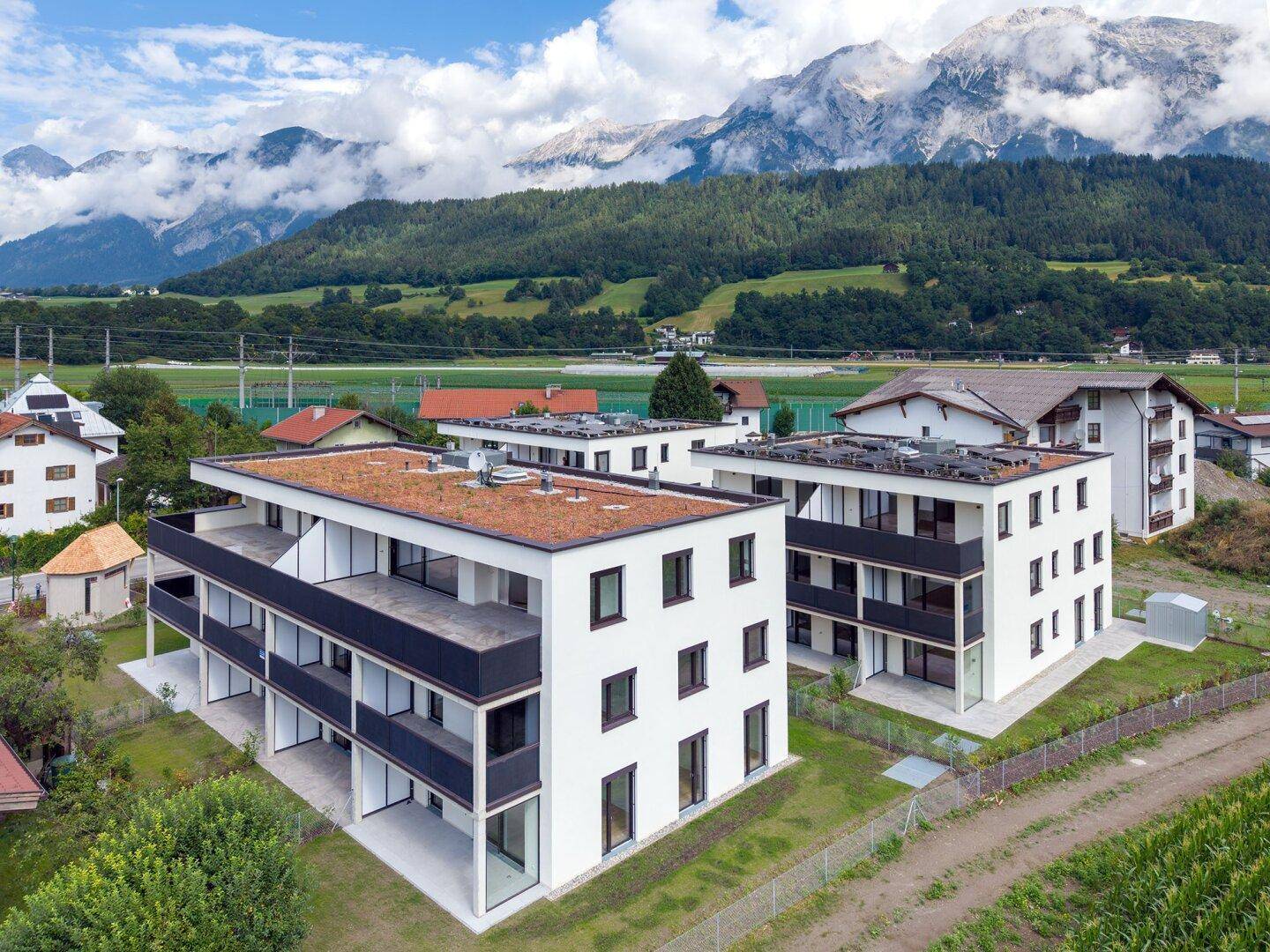 Moderne Mehrfamilienhäuser mit Flachdächern und Balkonen, umgeben von grüner Landschaft und Bergen.