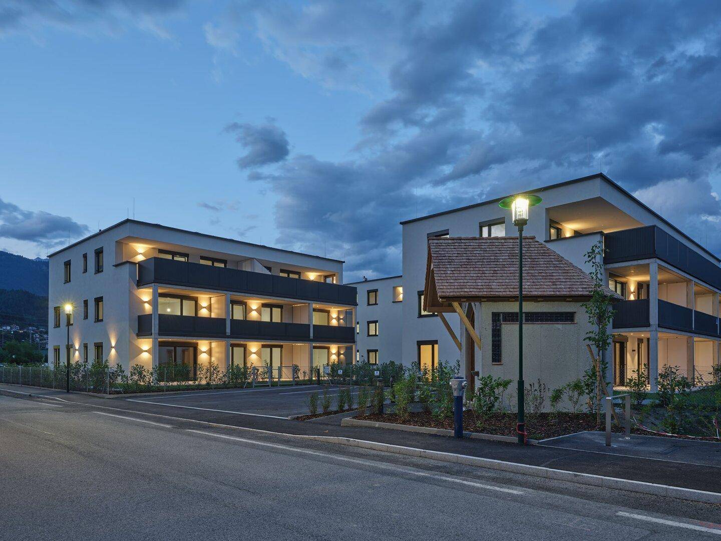 Moderne Wohngebäude bei Dämmerung mit beleuchteten Balkonen und Straßenbeleuchtung.