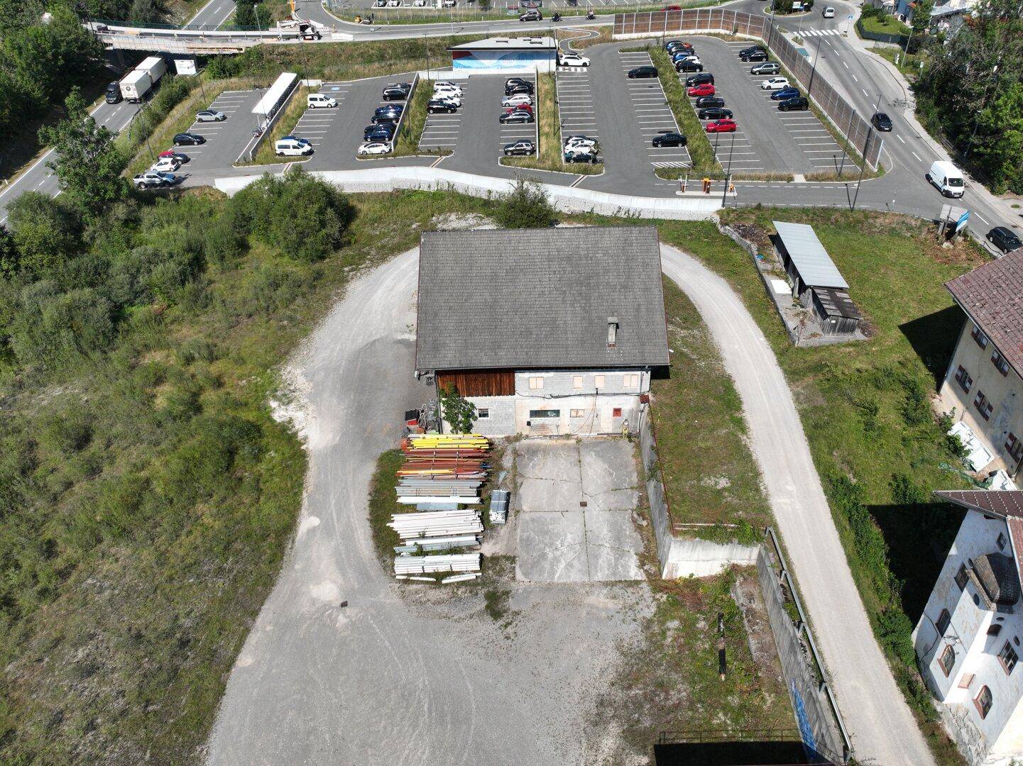 Nahaufnahme eines Scheunengebäudes mit unbefestigtem Vorplatz und einem großen Parkplatz im Hintergrund.