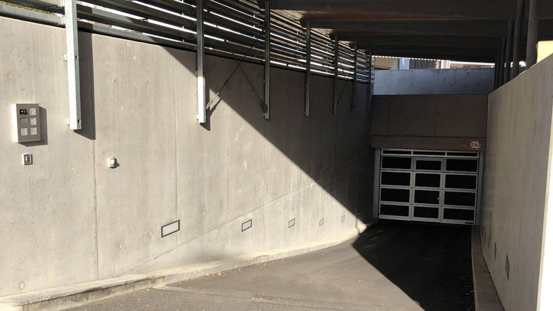 Einfahrt zur Tiefgarage mit Betonwänden, Metalltor und Gegensprechanlage im Sonnenlicht.