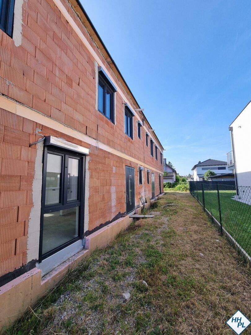 Längsseite des Neubaus im Rohbau mit Ziegelwänden und Fenstern, angrenzend an einen schmalen Grünstreifen.