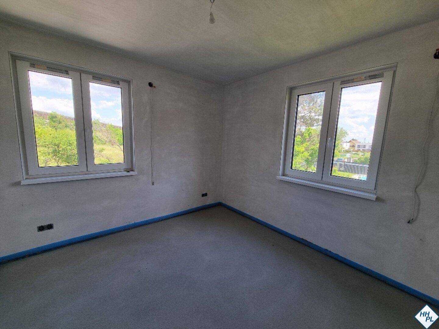 Unfertiges Zimmer mit Estrichboden und zwei Fenstern, die einen Blick ins Grüne bieten.