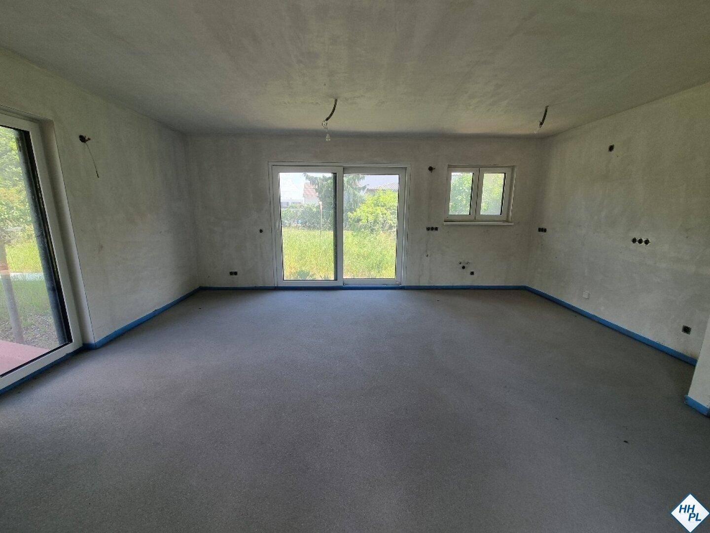 Unfertiger Wohnbereich mit Estrichboden, großen Schiebetüren und einem Fenster zum Garten.