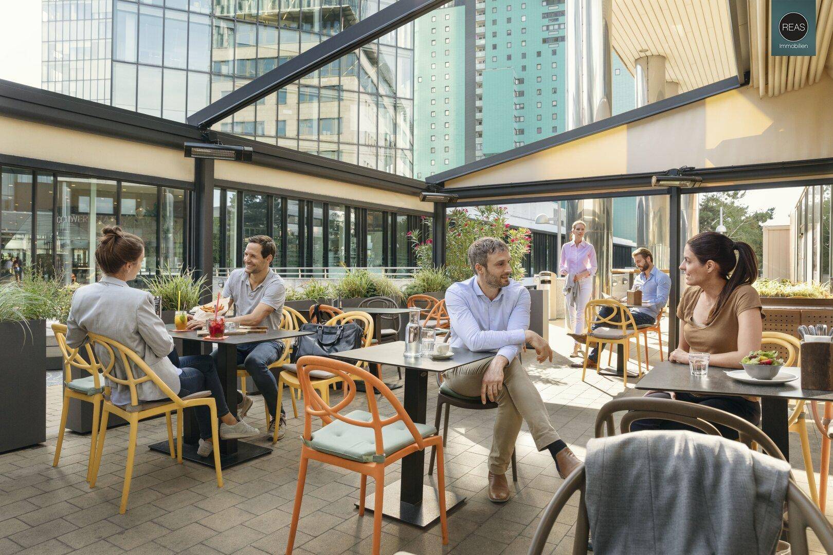 Belebtes Café auf einer Außenterrasse mit modernen Möbeln und Blick auf Gebäude.