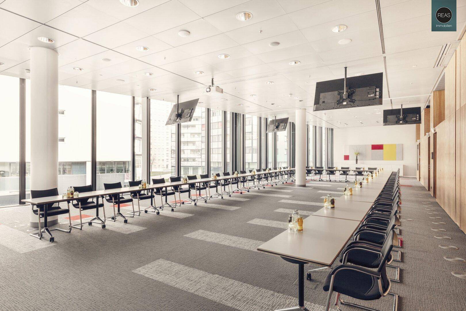Großer Konferenzraum mit Reihen von Tischen, Stühlen und mehreren Bildschirmen.