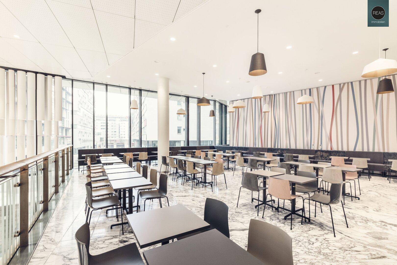 Helle Cafeteria mit vielen Tischen und Stühlen, umgeben von großen Fenstern.
