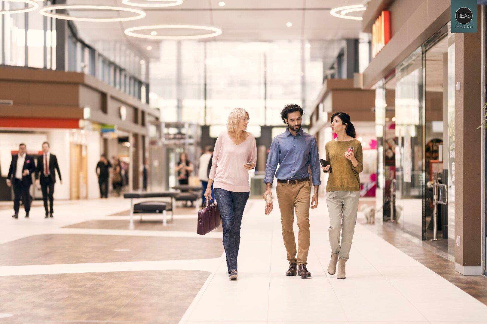 Menschen gehen durch eine helle, moderne Ladenpassage mit großen Schaufenstern.