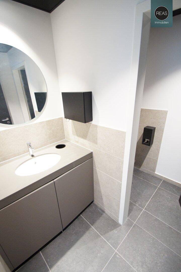 Modernes Badezimmer mit Waschbecken, Spiegel und grauen Fliesen, schlicht und funktional gestaltet.