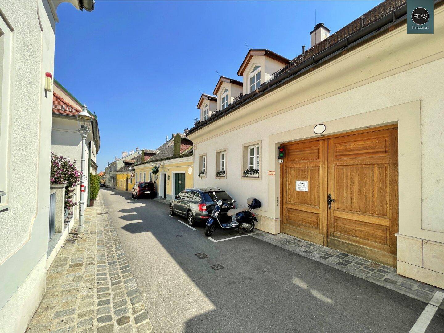 Enge Gasse mit traditionellen Gebäuden, gepflastertem Gehweg und parkenden Fahrzeugen an einem sonnigen Tag.