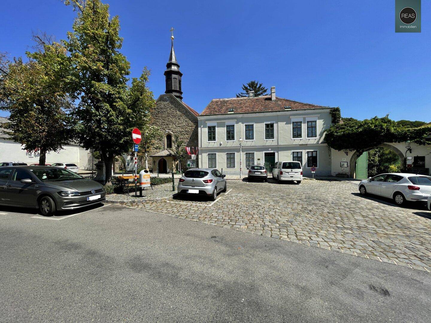 Historischer Dorfplatz mit Kirche, alten Gebäuden und gepflastertem Bereich unter blauem Himmel.