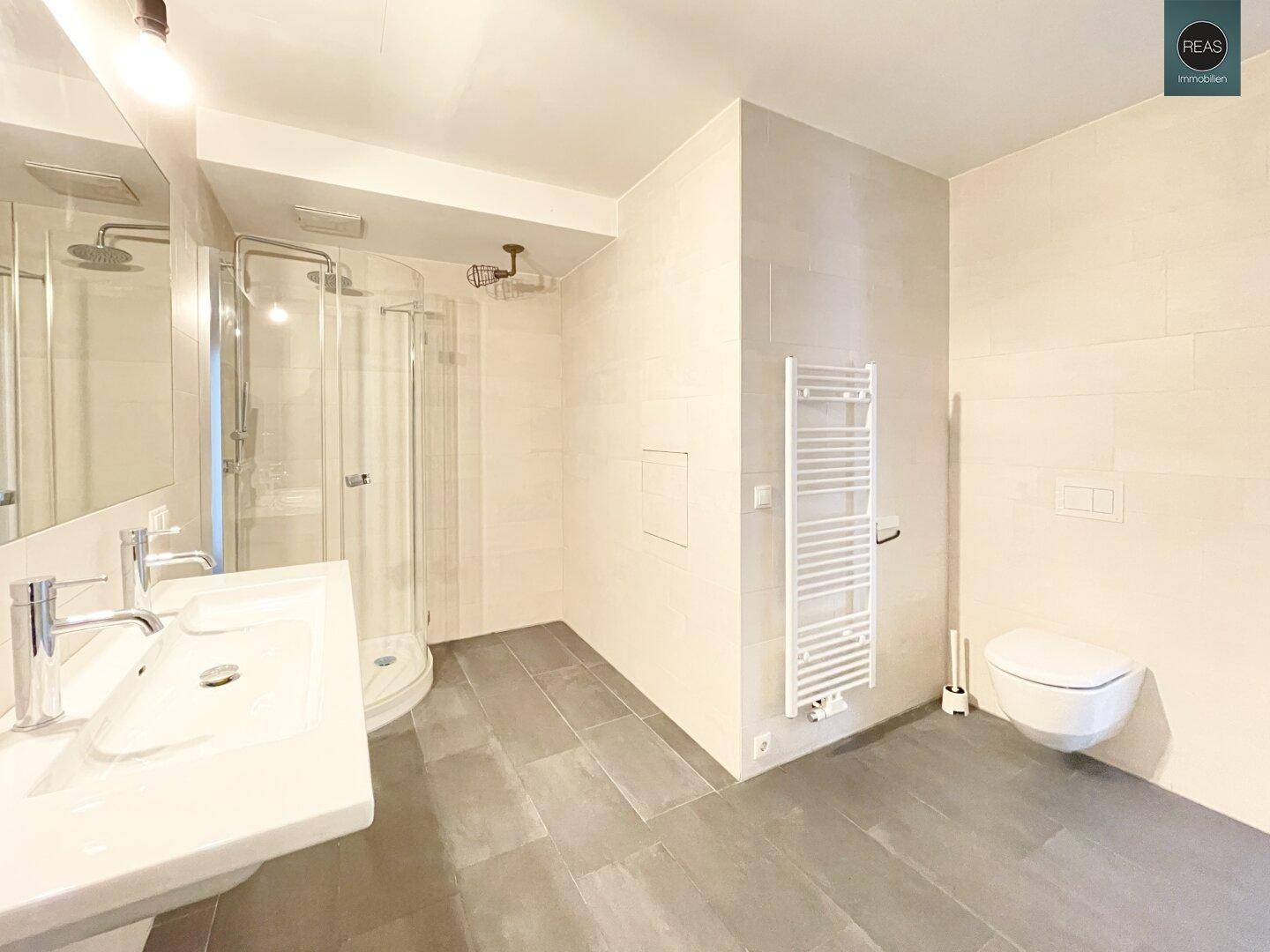 Modernes Badezimmer mit Duschkabine, wandhängendem WC und Doppelwaschbecken, in hellen Fliesen gehalten.