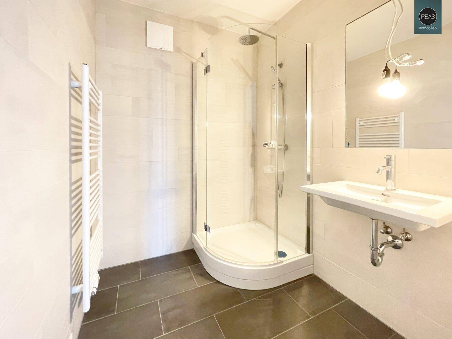 Modernes Badezimmer mit Duschkabine, Waschbecken und Handtuchheizkörper, in hellen Fliesen gehalten.
