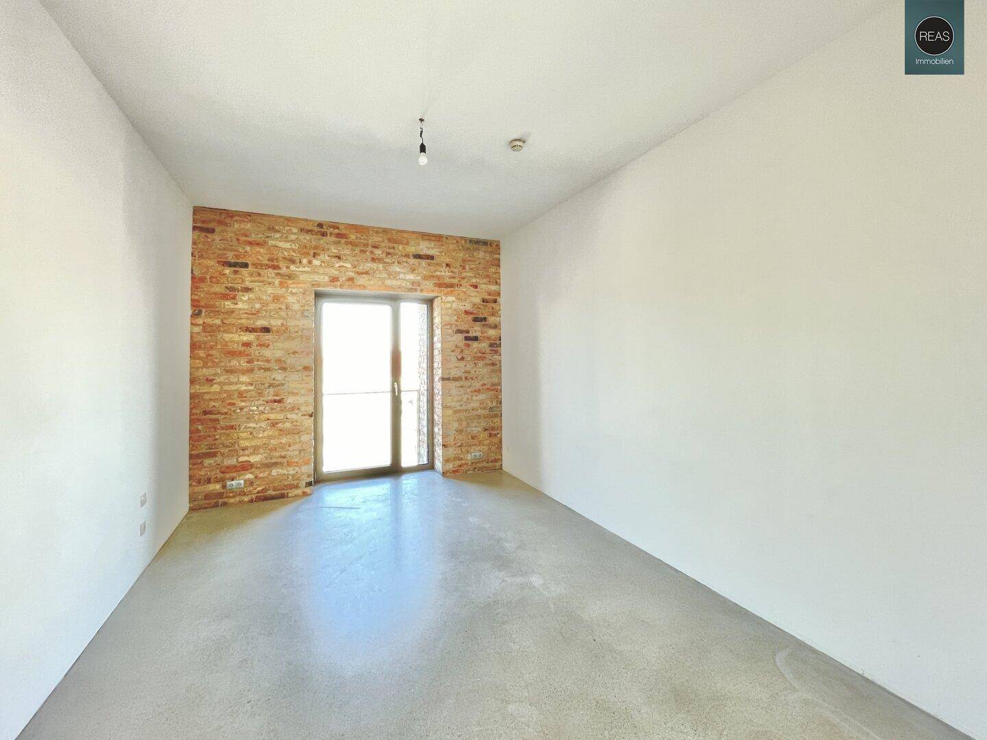 Heller, leerer Raum mit unverputzter Ziegelwand und einer Tür, die auf einen Balkon führt.