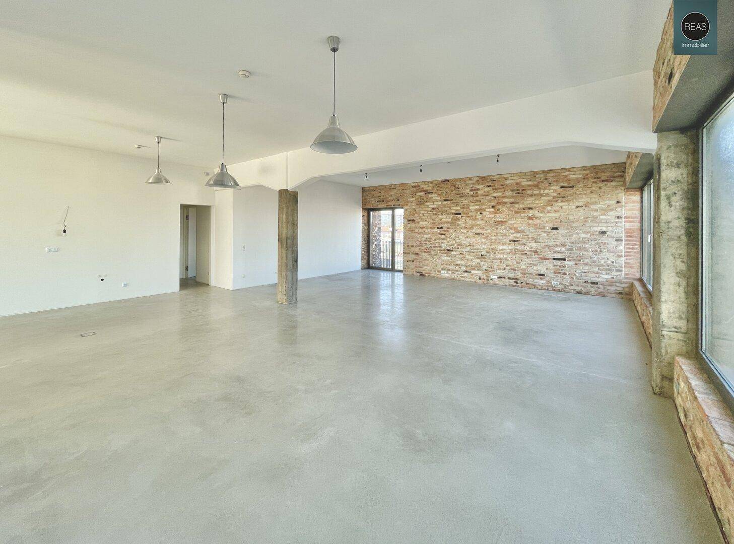 Geräumiger, leerer Büroraum mit Betonboden, unverputzten Ziegelwänden und großen Fenstern, die viel Tageslicht hereinlassen.