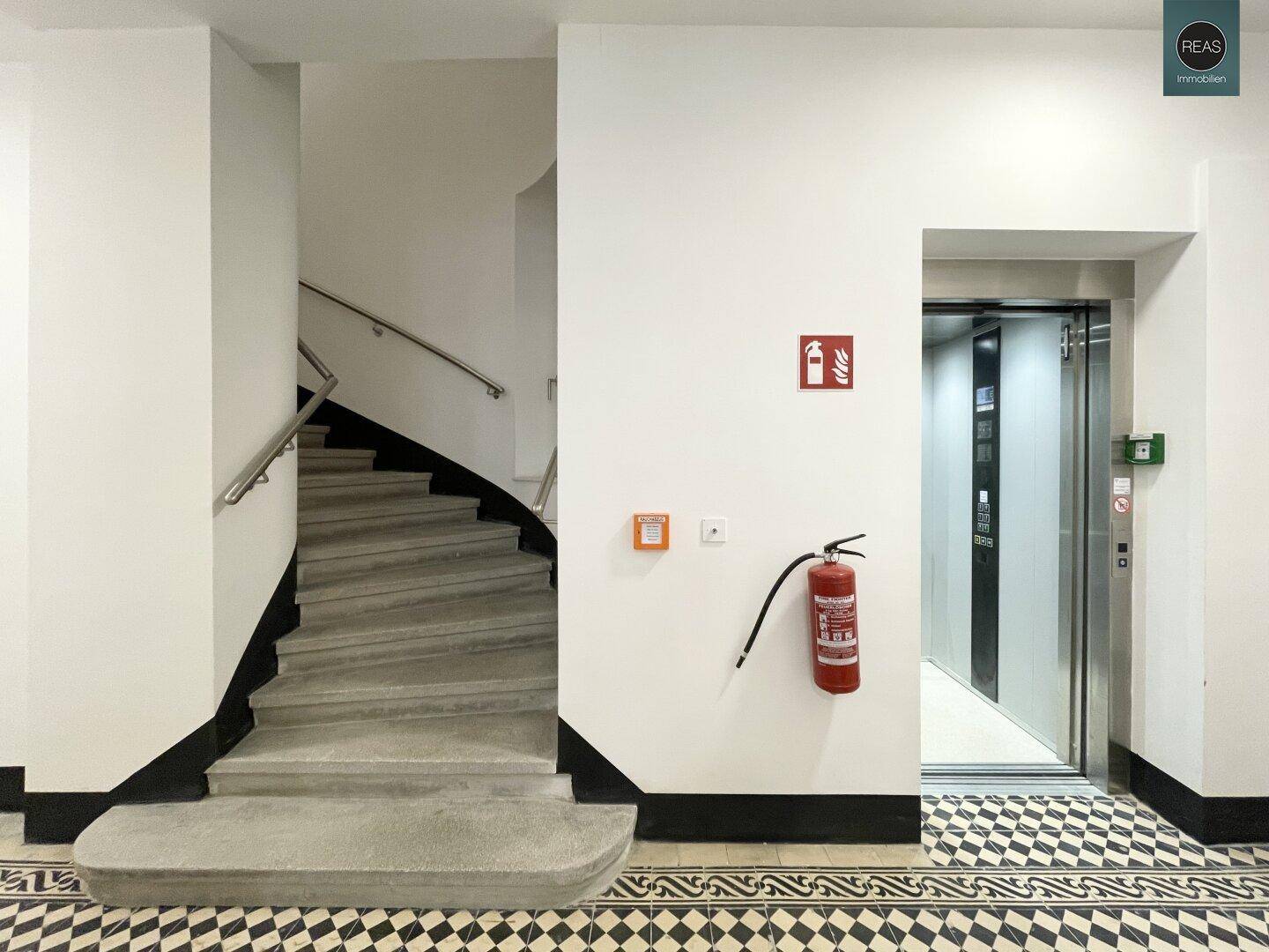 Treppenhaus und moderner Aufzug in einem Gebäude mit gemustertem Fliesenboden und Brandschutzvorrichtung.