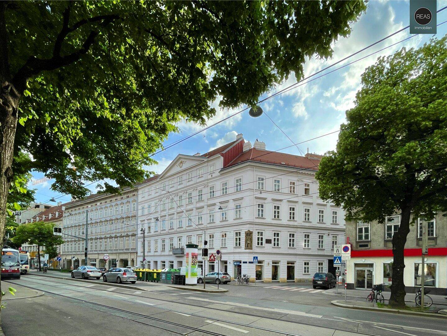 Historisches Gebäude an einer belebten Straße mit Bäumen und Straßenbahnschienen.