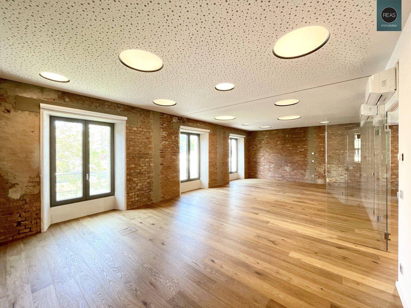 Geräumiger, moderner Büroraum mit freigelegten Ziegelwänden, Holzboden und vielen Einbauleuchten.