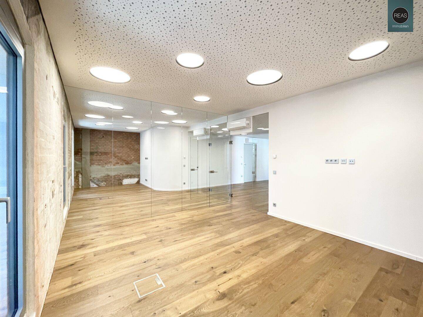 Moderner Büroraum mit Holzboden, weißen Wänden und Glasabtrennungen, die eine offene Atmosphäre schaffen.
