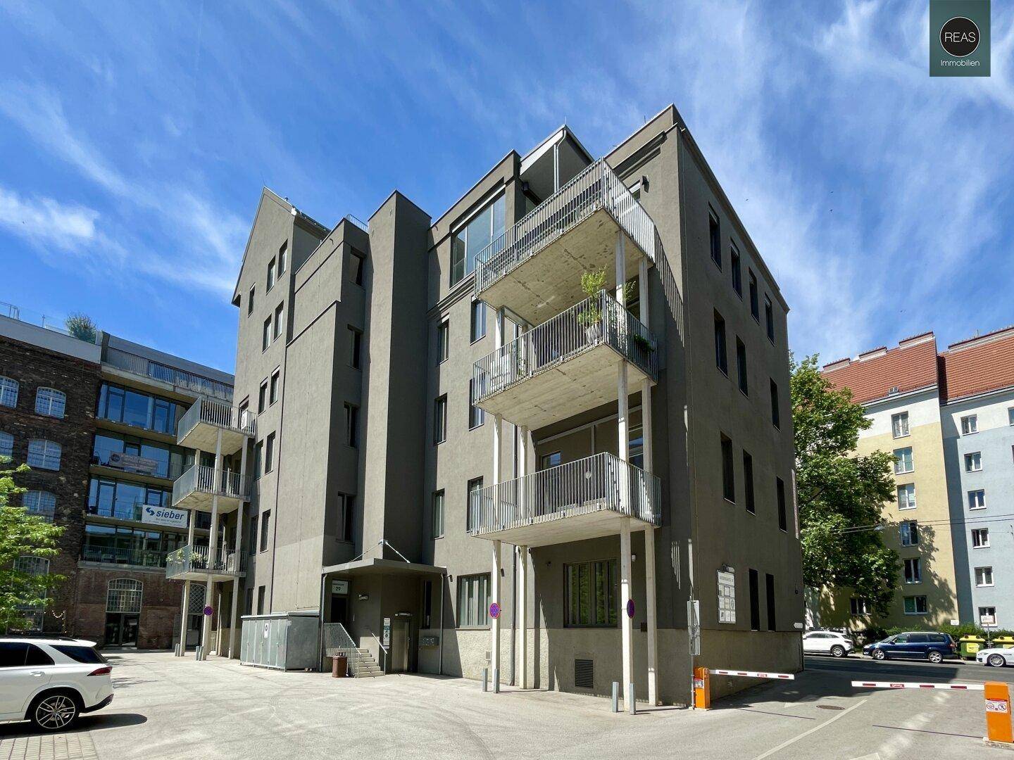 Modernes Gebäude mit grauer Fassade und mehreren Balkonen, umgeben von Parkplätzen.