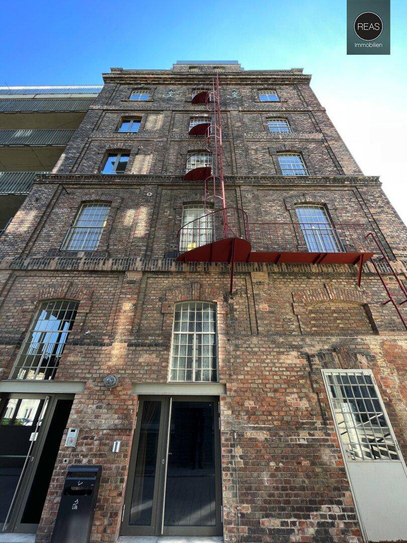 Hohes Backsteingebäude mit vielen Fenstern und einer roten Feuerleiter an der Fassade unter blauem Himmel.