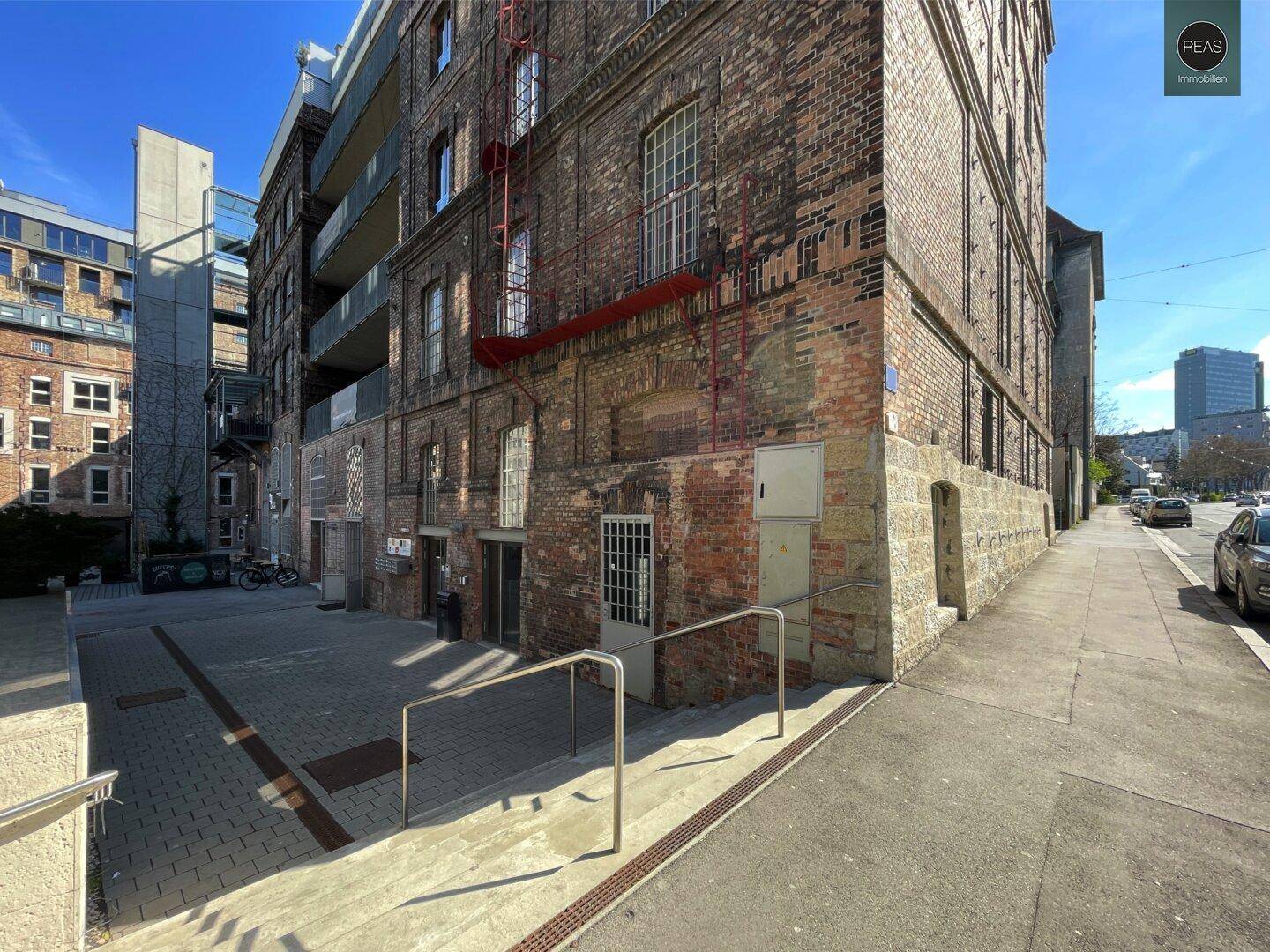 Eckansicht des Backsteingebäudes mit gepflastertem Gehweg, Treppenaufgang und angrenzender Straße.