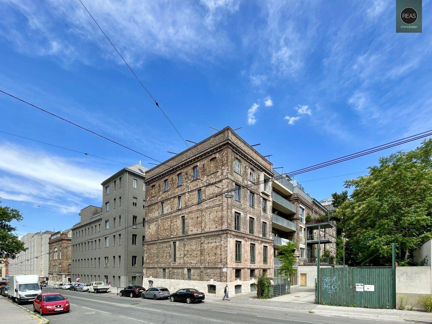 Weitläufige Straßenansicht mit dem Backsteingebäude, modernen Nachbargebäuden und parkenden Autos unter blauem Himmel.