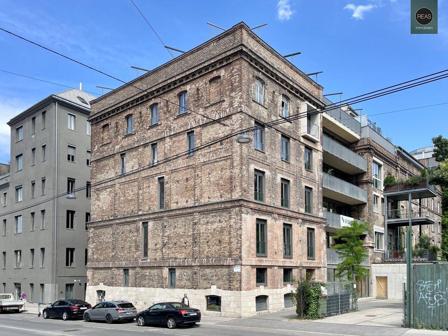 Schräge Ansicht des Backsteingebäudes mit modernen Anbauten, Balkonen und geparkten Fahrzeugen an der Straße.