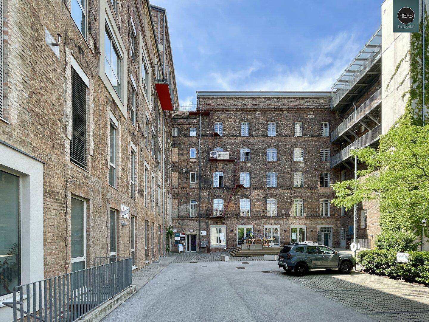 Innenhof zwischen zwei Backsteingebäuden mit einem geparkten Auto und viel Grün an einem sonnigen Tag.