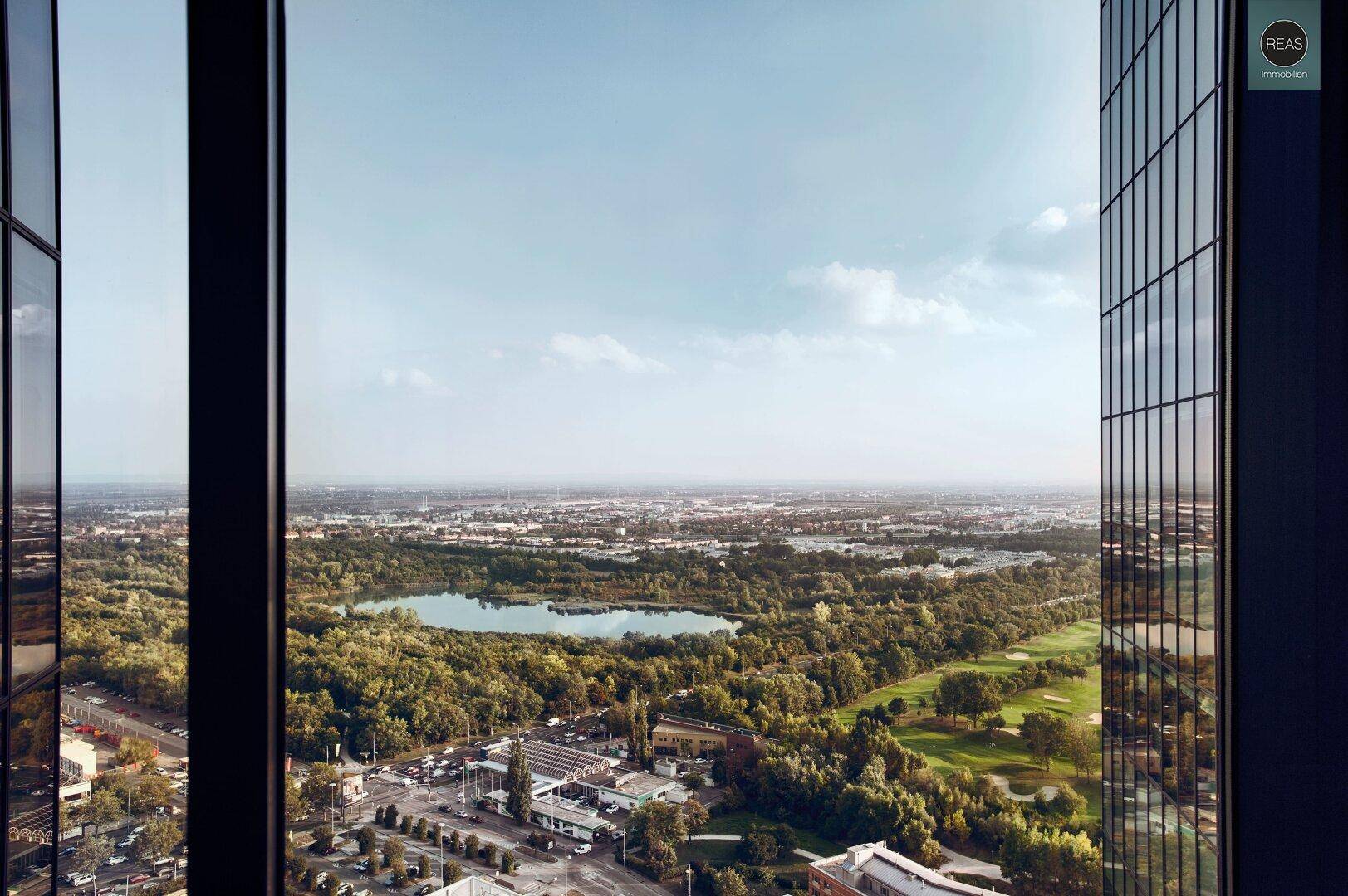 Panoramablick aus einem Hochhausfenster auf eine grüne Landschaft mit See und Stadtgebiet.