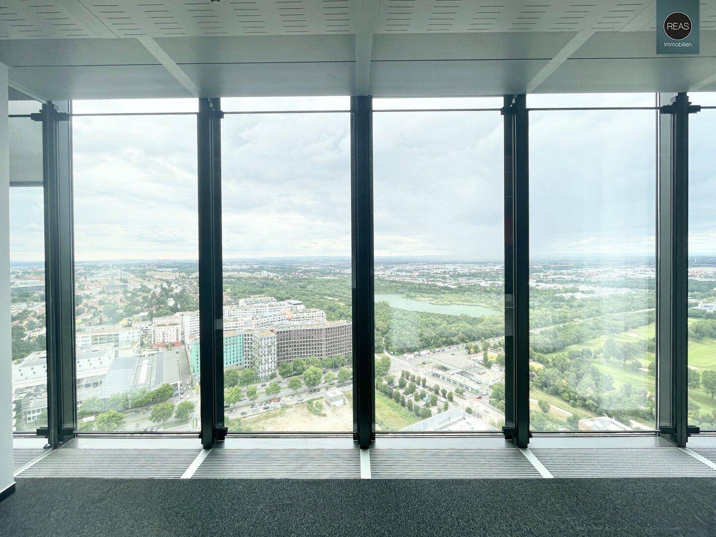 Weitblick aus dem Bürofenster auf die Stadt und umliegende Grünflächen unter bewölktem Himmel.