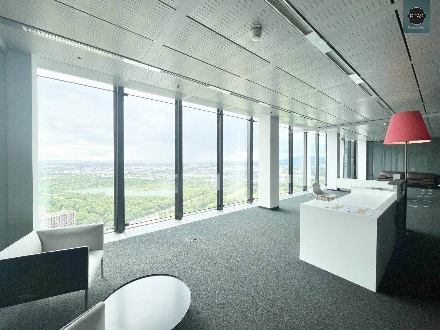Empfangsbereich oder Lounge im Büro mit modernen Möbeln und einer roten Stehlampe, umgeben von großen Fenstern.