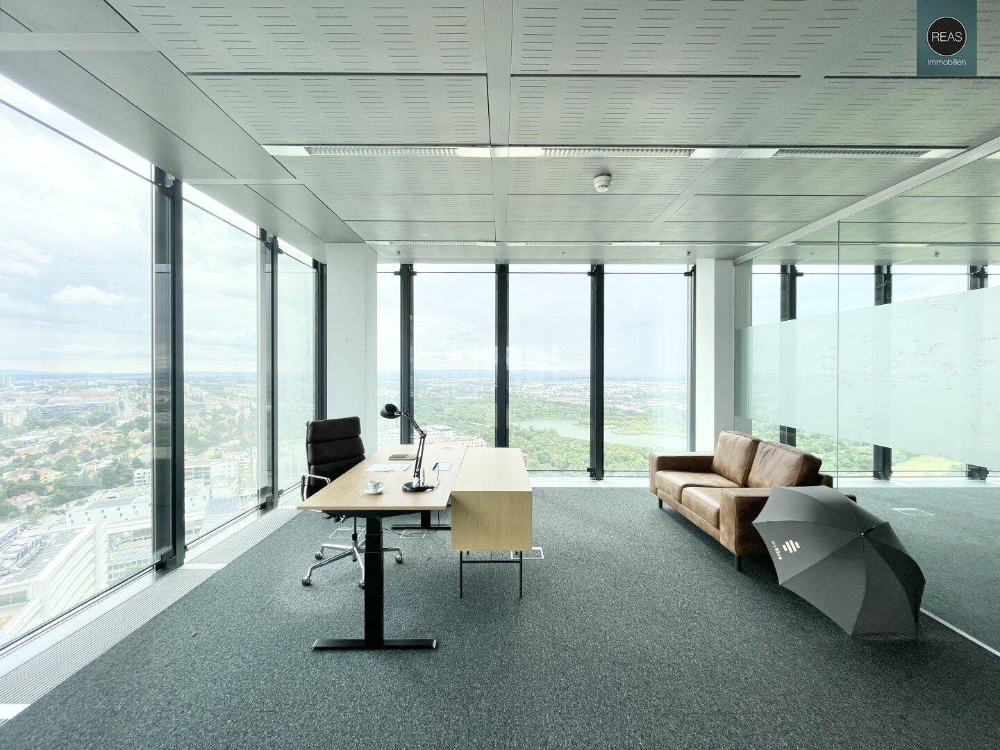 Helles Einzelbüro mit Schreibtisch, Ledersofa und weitem Blick über die Stadt und Grünflächen.