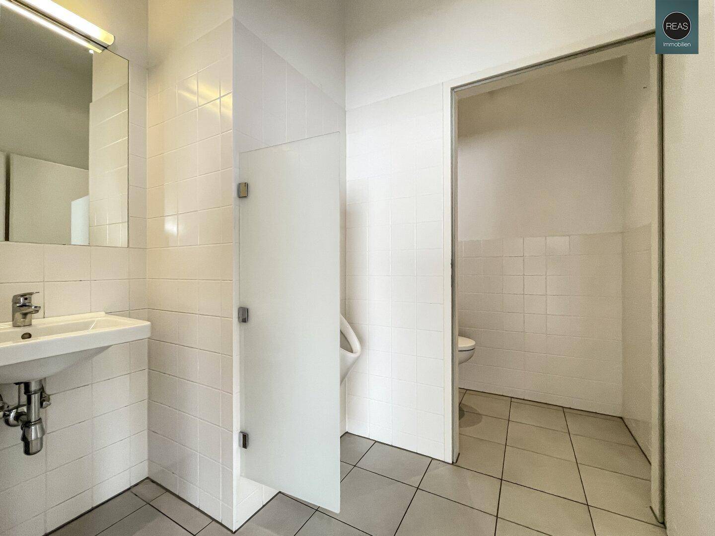 Saubere Toilettenanlage mit weißen Fliesen, Waschbecken und Urinal in modernem Design.