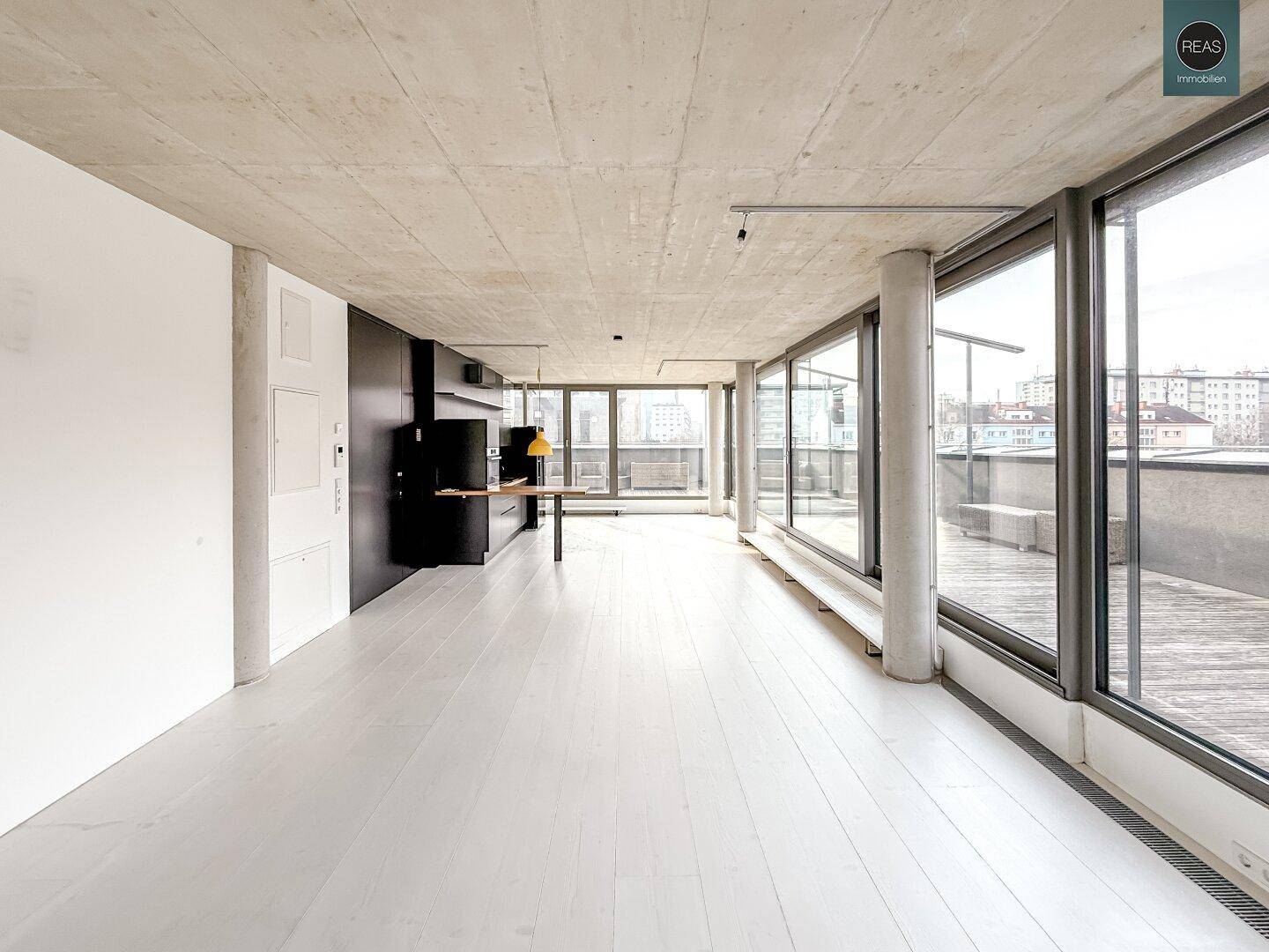 Langer, heller Raum mit Holzboden, Sichtbetondecke und großen Fenstern zur Dachterrasse.