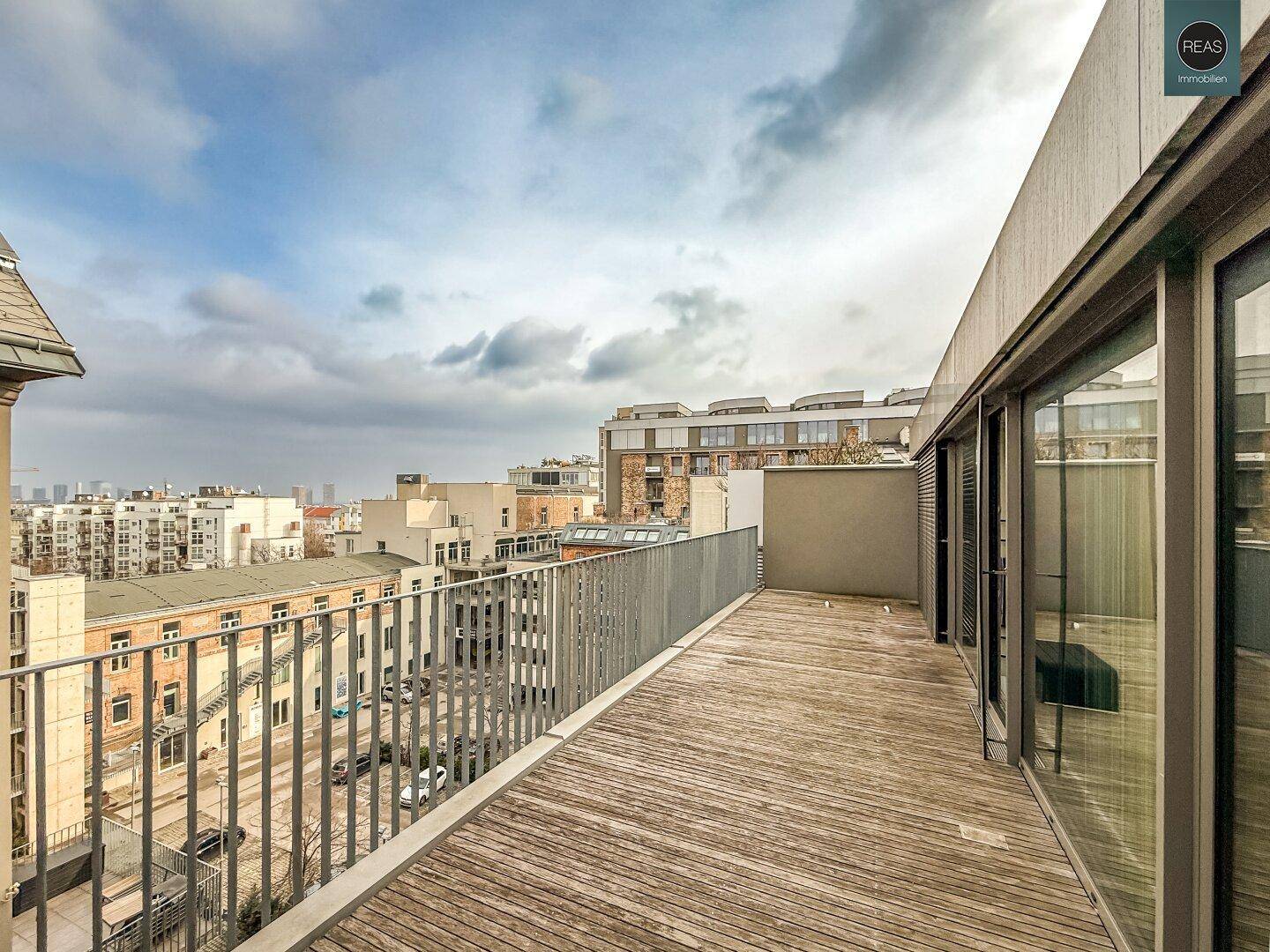 Lange Dachterrasse mit Holzboden und Metallgeländer, die einen weiten Stadtblick über die Dächer ermöglicht.