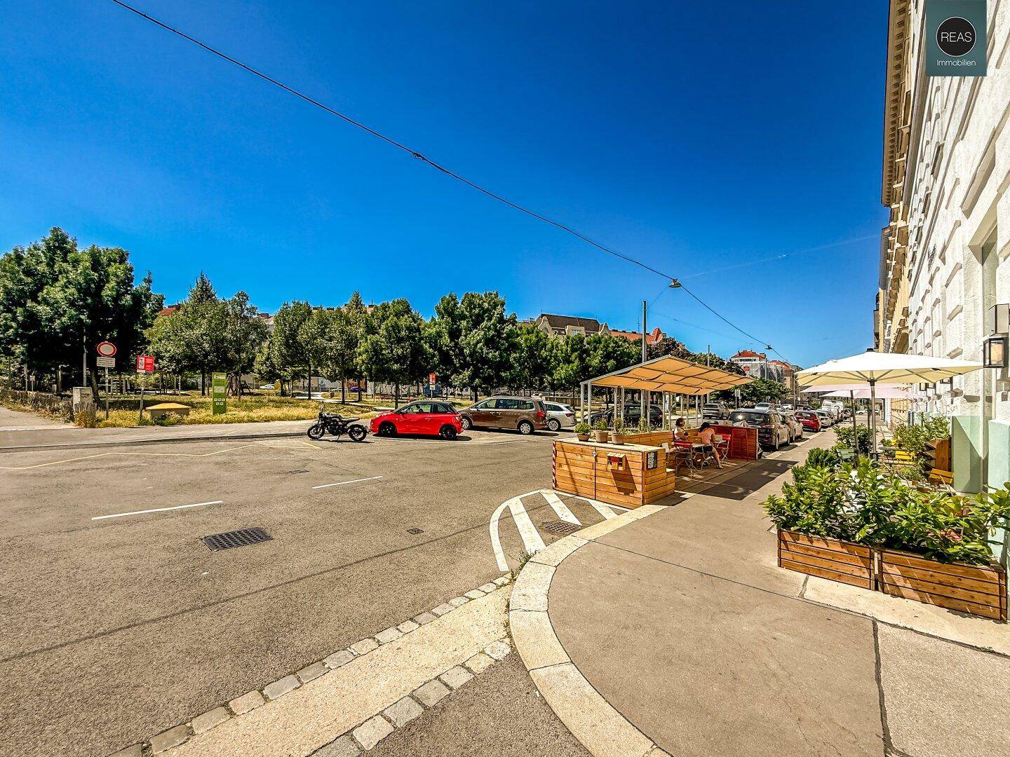 Belebtes Stadtviertel mit Bäumen, Cafés und geparkten Fahrzeugen an einem sonnigen Tag.