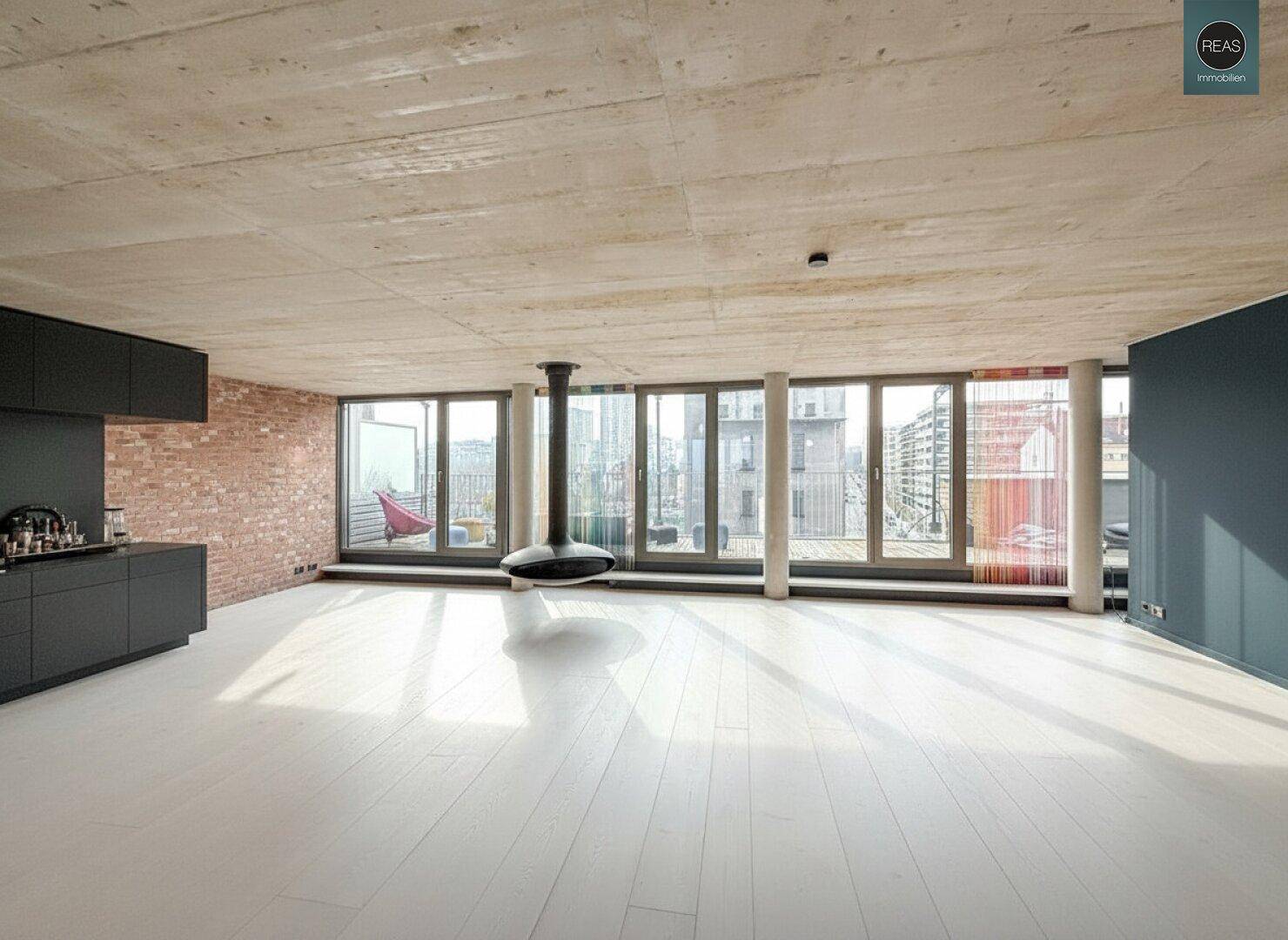 Heller, offener Raum mit weißem Holzboden, Sichtbetondecke und einem modernen Hängekamin als Blickfang.