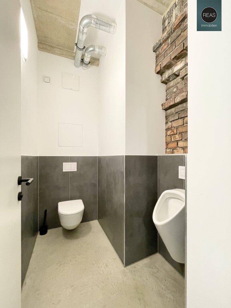 Separate Toilette mit Urinal, Wand-WC und freiliegenden Rohren an der Decke, im industriellen Stil.